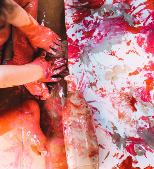 Little girl makes a mess with paints on a white canvas on the floor of her house.
