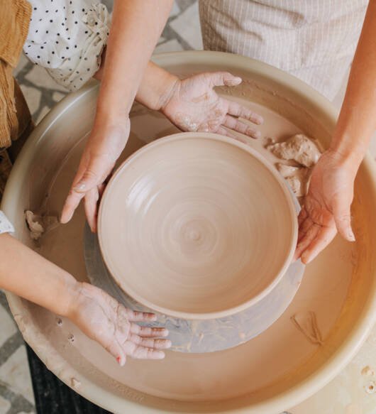 Top view of two children shaping a ceramic bowl on a pottery wheel. Hands only, no faces.