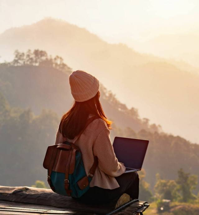 Young woman freelancer traveler working online using laptop and enjoying the beautiful nature landscape with mountain view at sunrise
